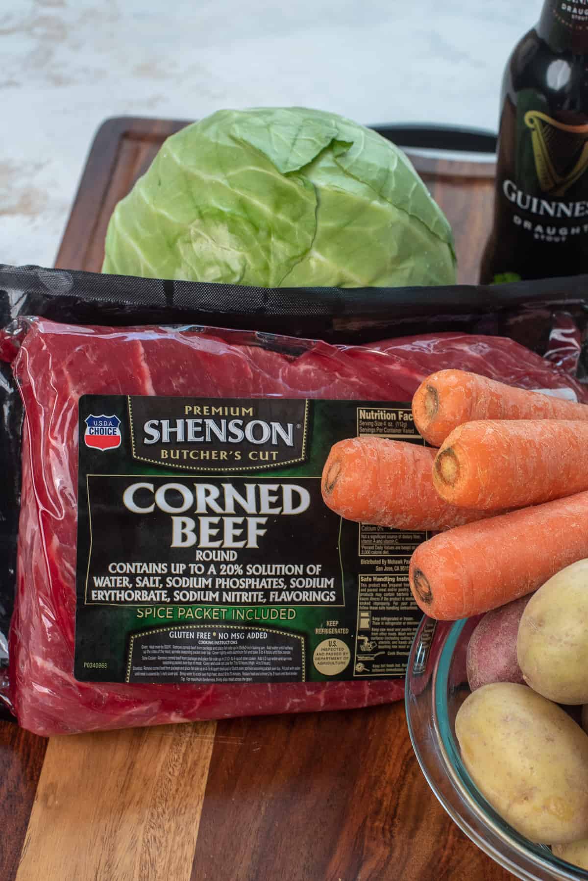 Ein Shenson Corned Beef auf einem Holzbrett mit einem Kohlkopf und einer Schüssel Kartoffeln und Karotten.