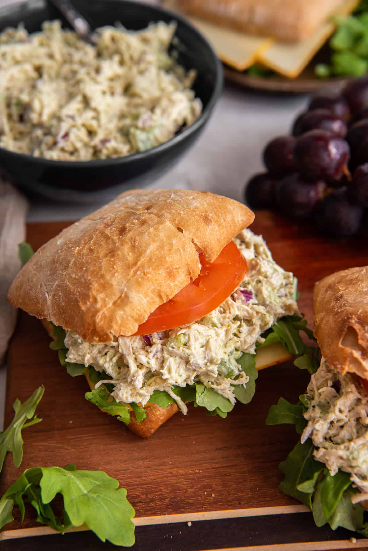 A pesto chicken salad sandwich on ciabatta bread with tomato and lettuce on a wood board.