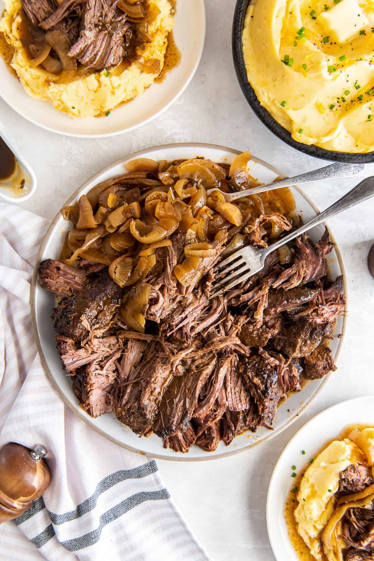 A top down shot of a plate of shredded green chile beef with onions next to a bowl of mashed potatoes.