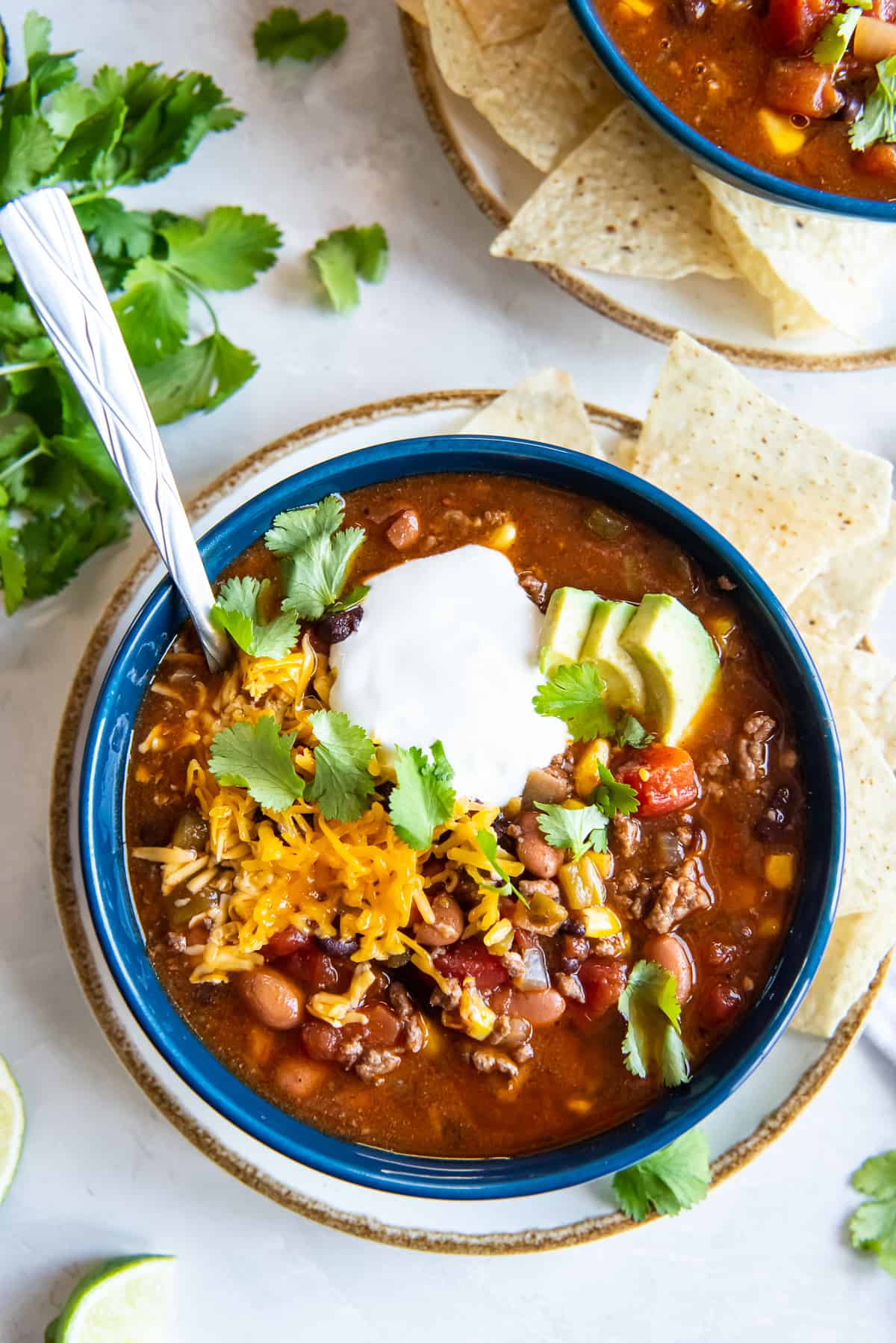 A top down shot of taco soup in a blue bowl with a spoon topped with sour cream, cilantro, and avocado.