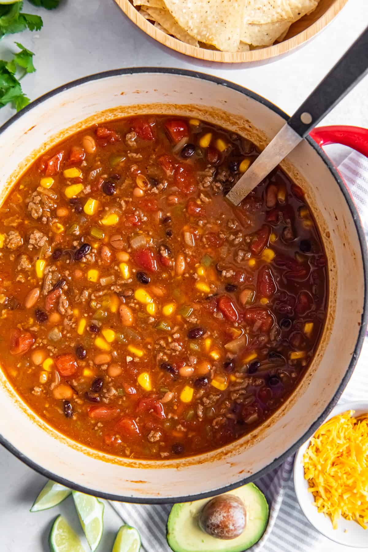 A top down shot of a ladle resting in a pot filled with taco soup with avocado and shredded cheese nearby.