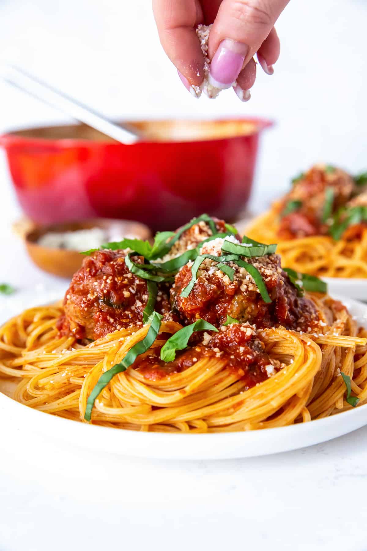 Fingers sprinkling Parmesan cheese on a plate of spaghetti and meatballs.