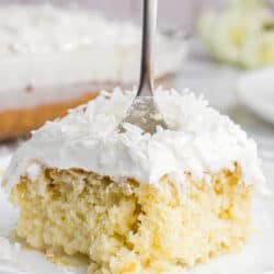 A slice of coconut poke cake on a white plate with a fork stuck in it.