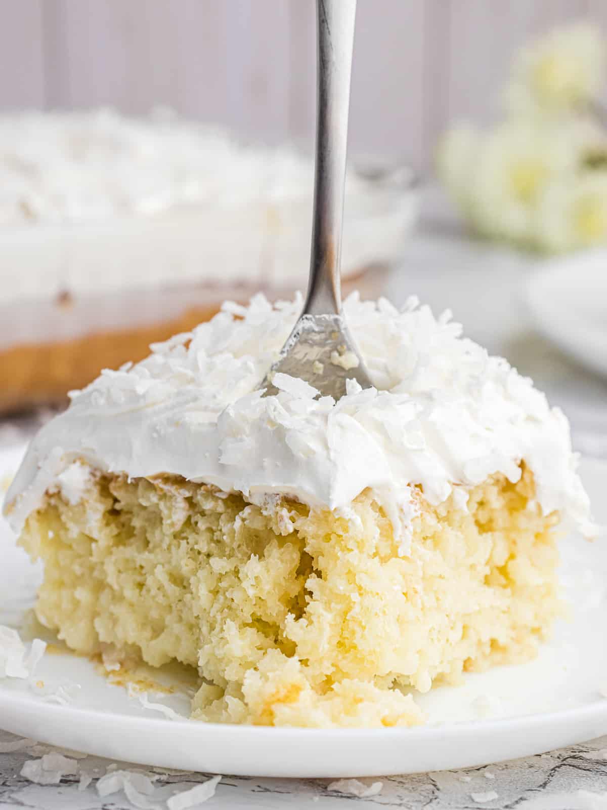 A slice of coconut poke cake on a white plate with a fork stuck in it.