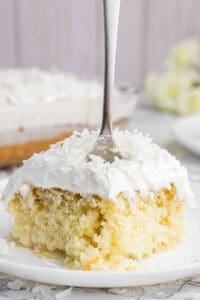 A slice of coconut poke cake on a white plate with a fork stuck in it.