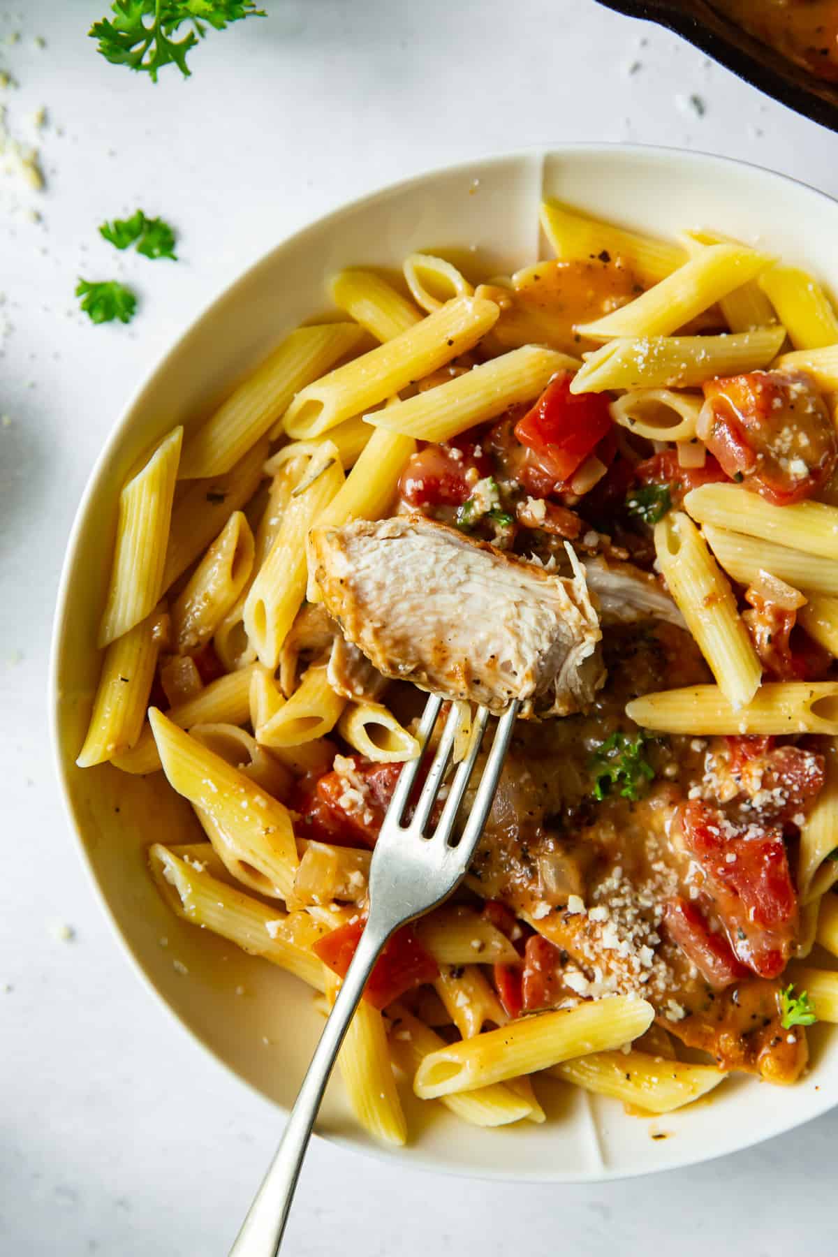 A fork piercing a piece of chicken in a bowl with pasta and tomatoes.