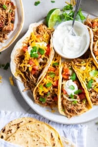A top down shot of a platter full of chicken tacos with a bowl of sour cream and bowls of other toppings nearby.