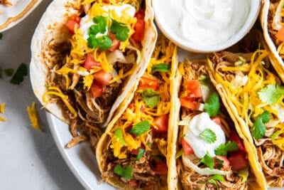 A top down shot of a platter full of chicken tacos with a bowl of sour cream and bowls of other toppings nearby.