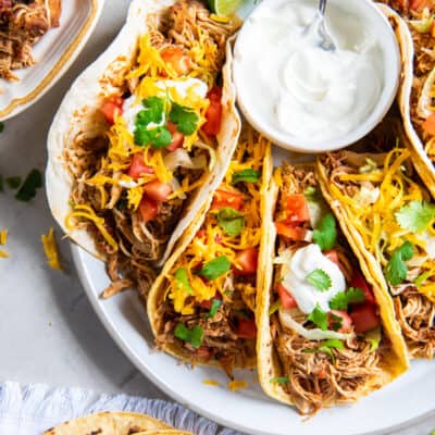 A top down shot of a platter full of chicken tacos with a bowl of sour cream and bowls of other toppings nearby.