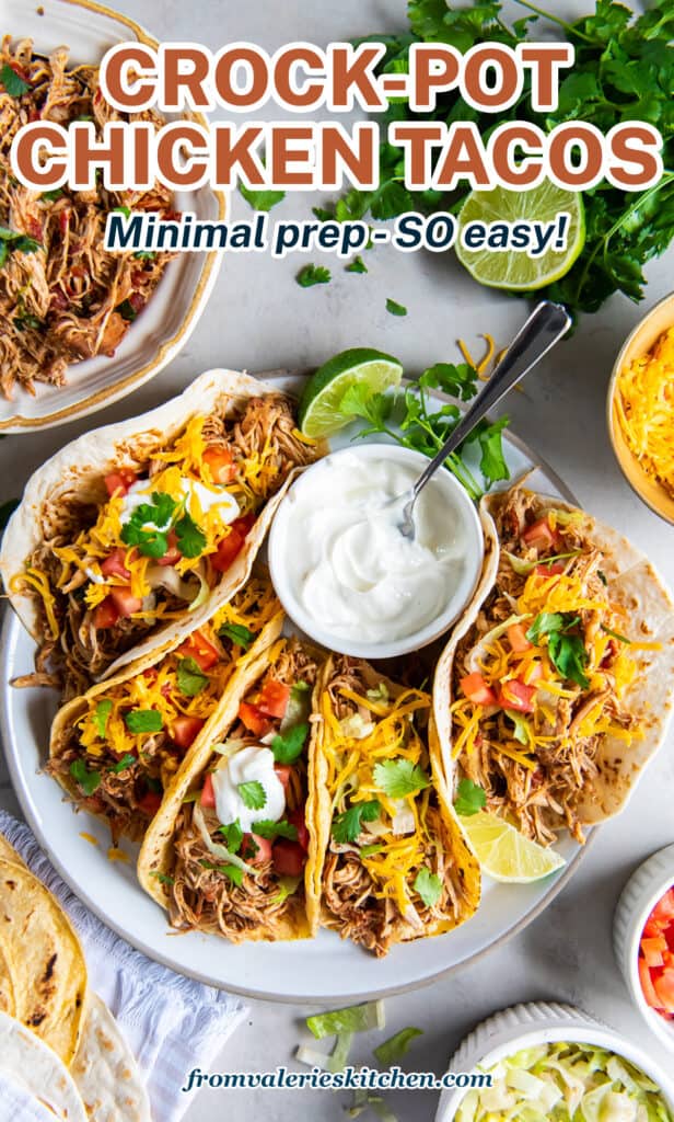 A top down shot of a platter full of chicken tacos with a bowl of sour cream and bowls of other toppings nearby with text.