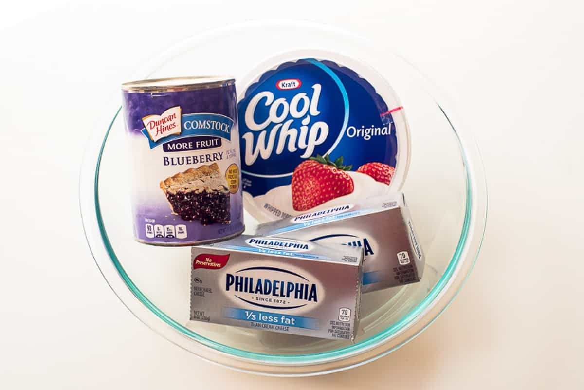 A can of blueberry pie filling, container of Cool Whip and package of cream cheese in a glass bowl.