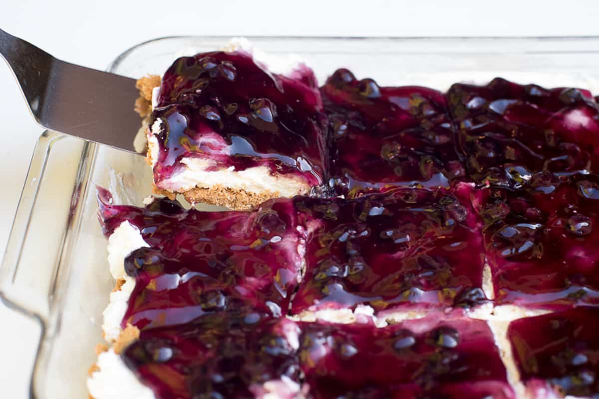 A spatula lifts a slice of blueberry cream cheese dessert from a baking dish.