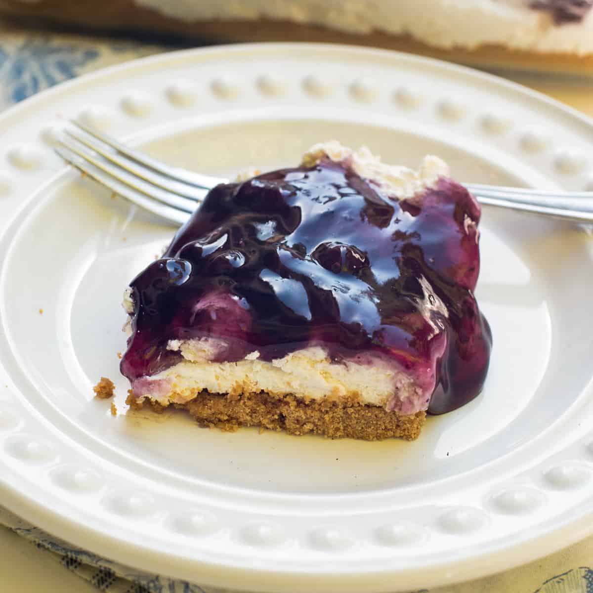 A piece of blueberry cheesecake dessert on a white plate with a fork.