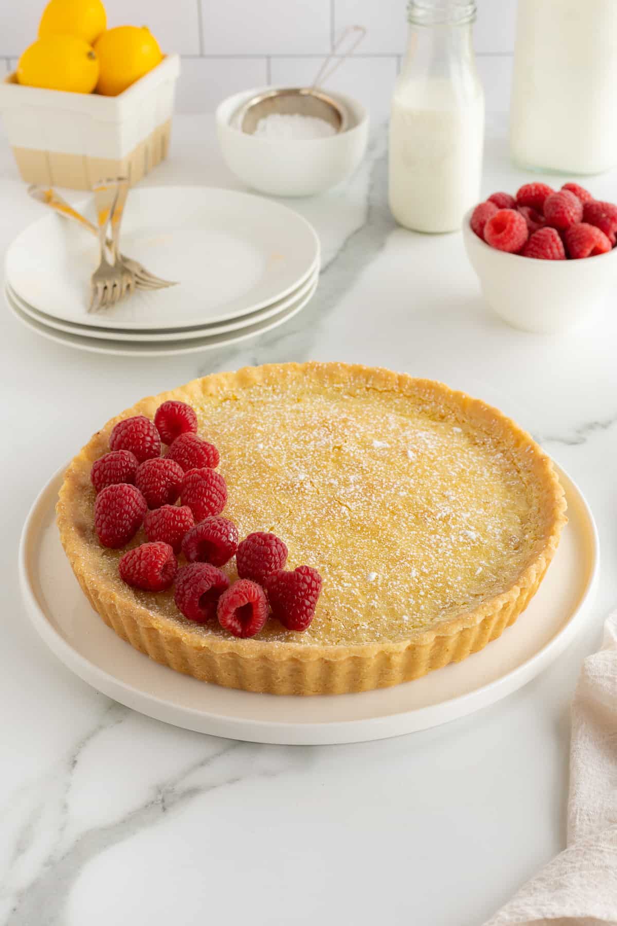 A lemon tart topped with raspberries in front of a stack of plates on a kitchen counter.