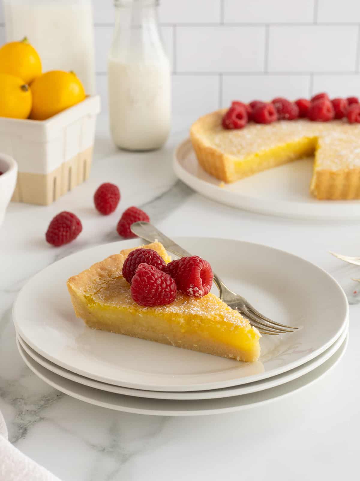 A slice of lemon tart topped with raspberries on a stack of white plates with a fork.