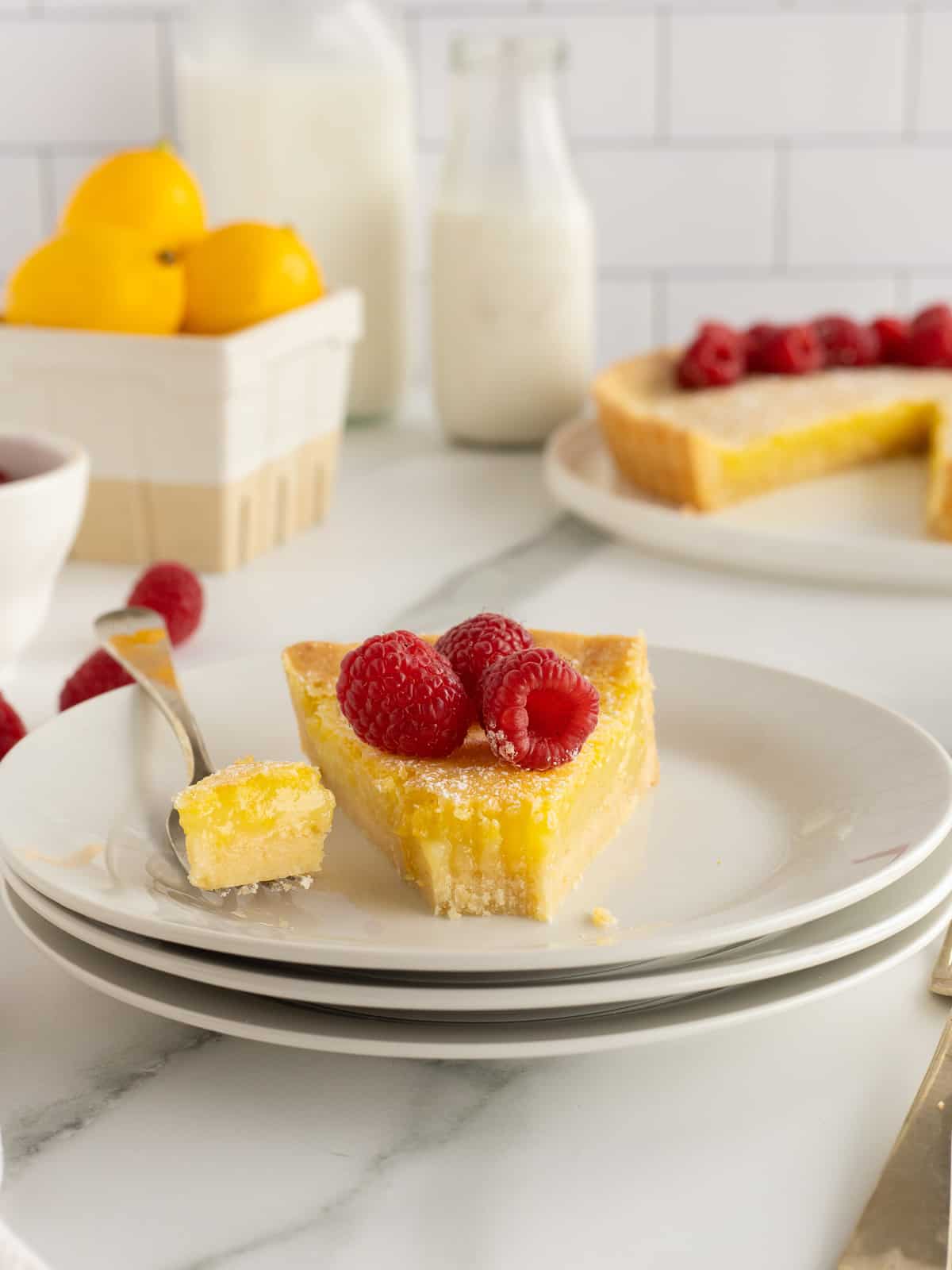 A slice of lemon tart topped with raspberries and a fork on a stack of white plates.