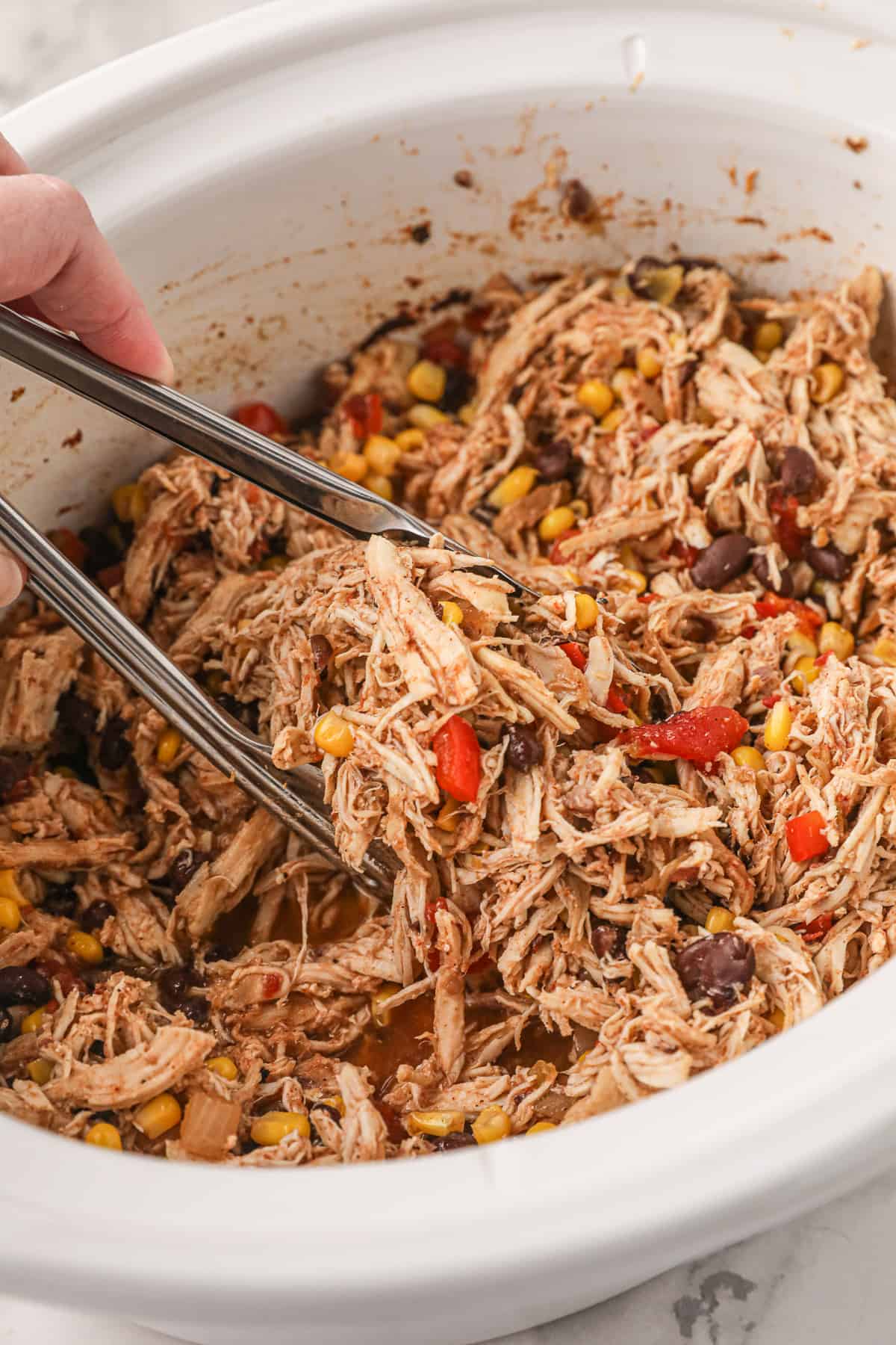 Metal tongs scooping shredded chicken from a white slow cooker.