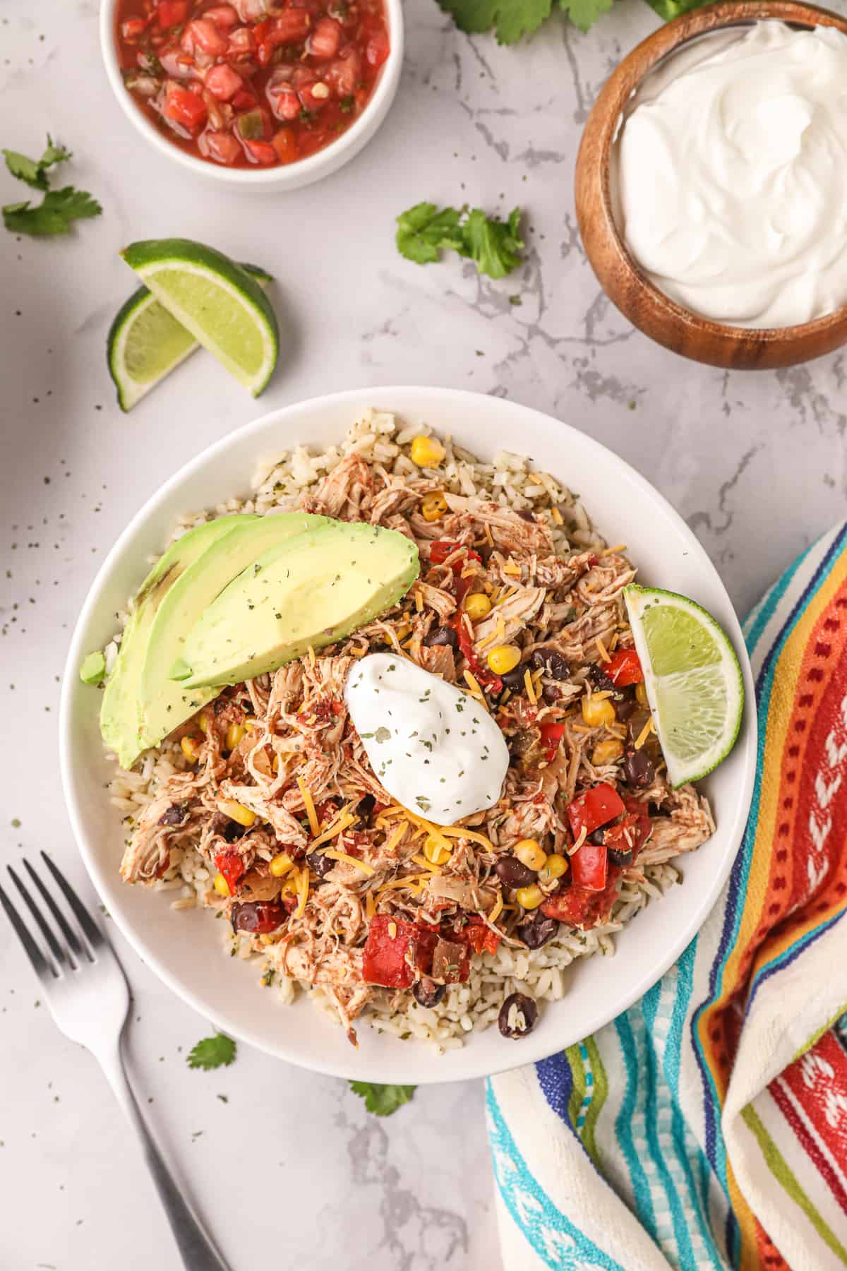 A top down shot of a bowl filled with shredded Tex-Mex Chicken over rice next to small bowls of salsa and sour cream.