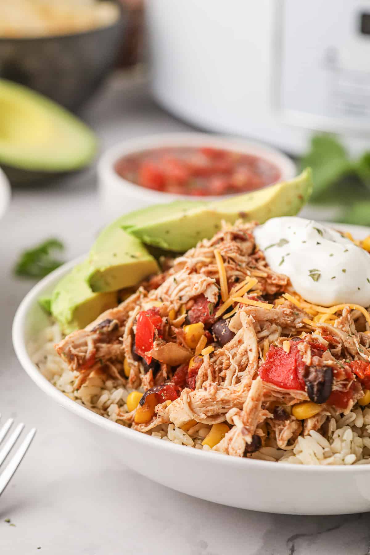 A closeup of a bowl of Tex-Mex chicken on rice with avocado and sour cream in front of a white slow cooker.