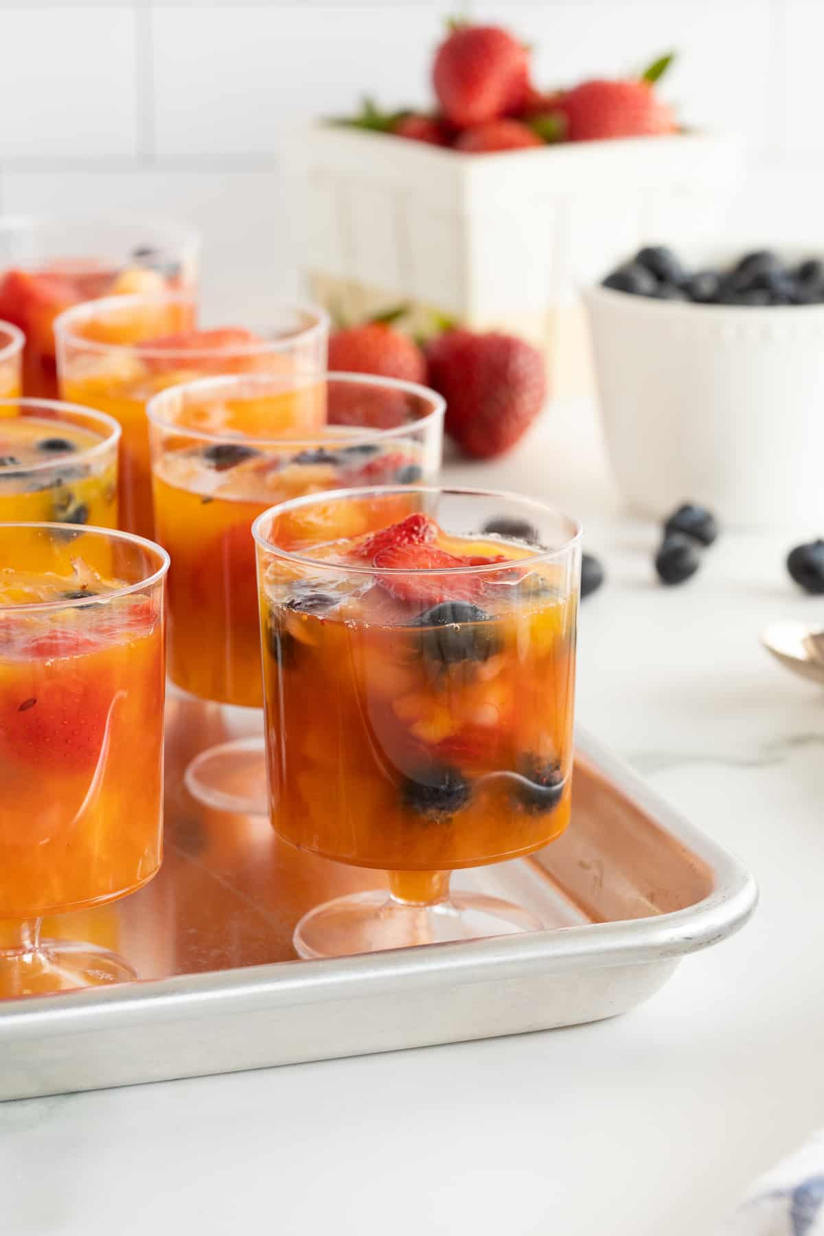 A close up of plastic cups filled with frozen fruit on a baking sheet next to bowls of strawberries and blueberries.