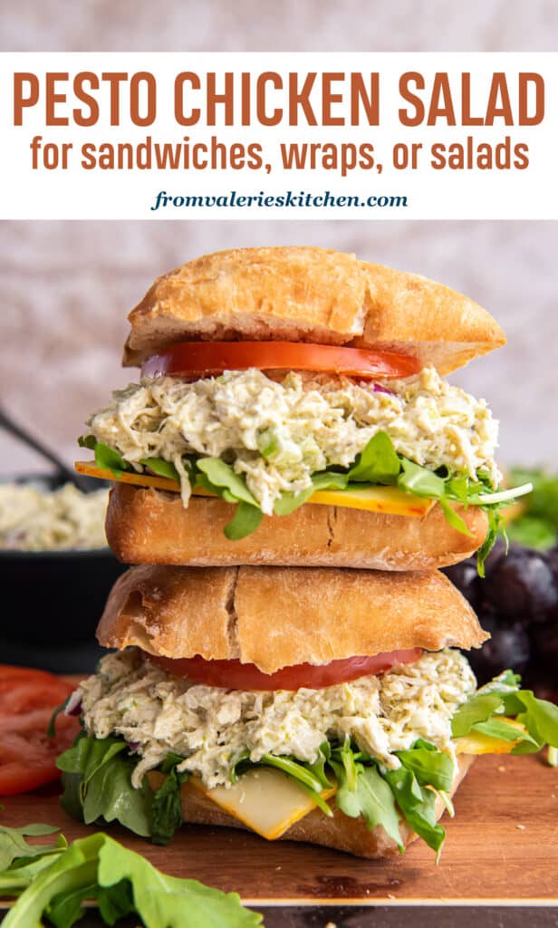 Two pesto chicken salad sandwiches on ciabatta with tomatoes and greens stacked on a wood board with text.