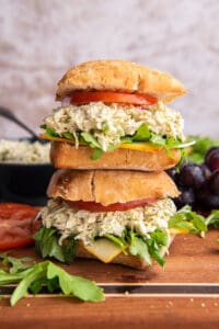 Two pesto chicken salad sandwiches on ciabatta with tomatoes and greens stacked on a wood board.