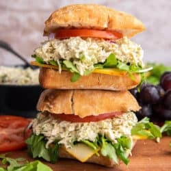 Two pesto chicken salad sandwiches on ciabatta with tomatoes and greens stacked on a wood board.