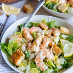 A fork resting in a bowl with caesar salad with shrimp and, croutons, and dressing.