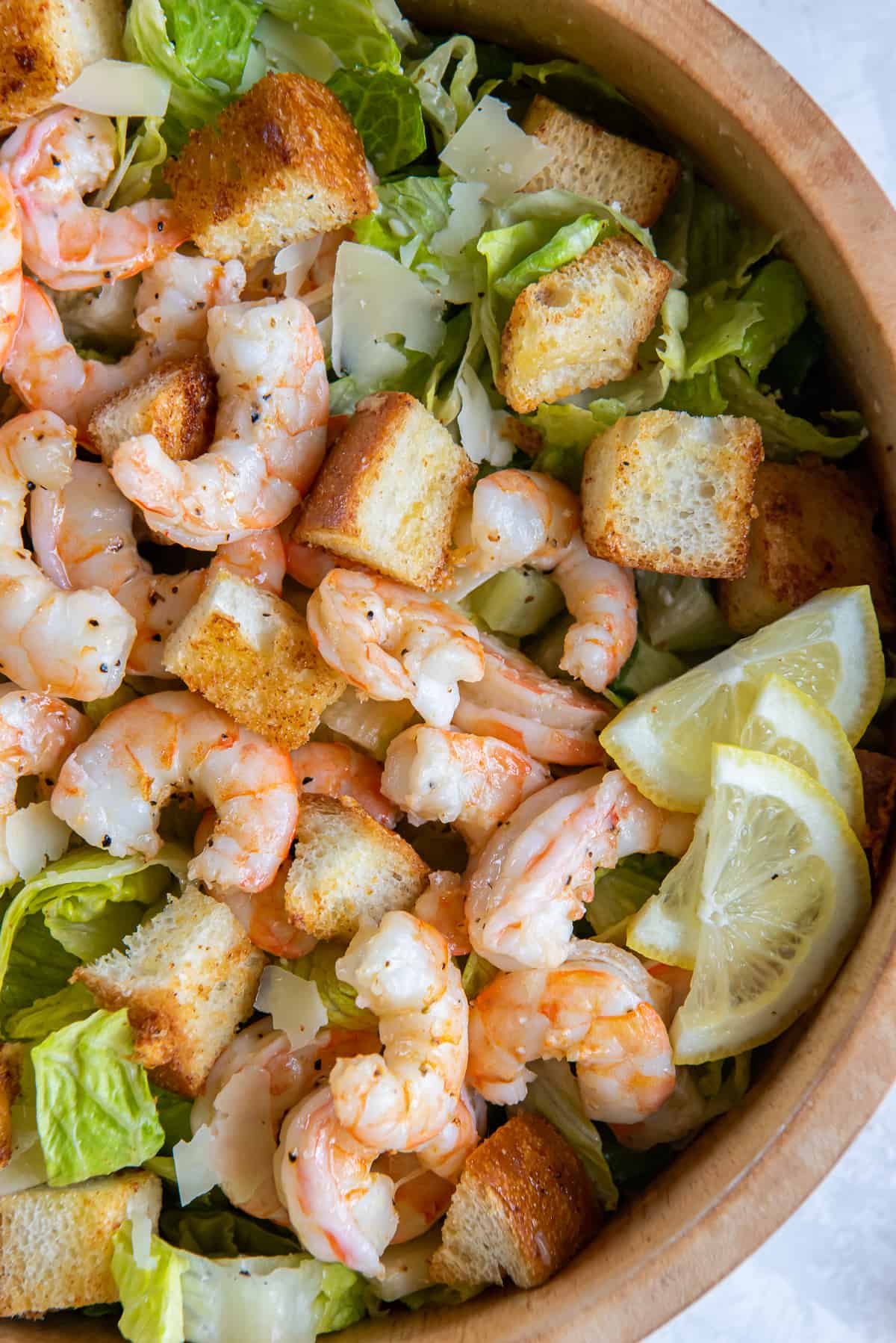A top down close up of roasted shrimp on a bed of romaine lettuce with croutons and slices of lemon in a wood bowl.