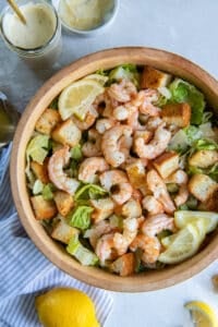 A top down shot of Shrimp Caesar Salad in a wood bowl near a mason jar filled with dressing and fresh lemons.