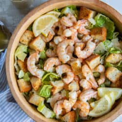 A top down shot of Shrimp Caesar Salad in a wood bowl near a mason jar filled with dressing and fresh lemons.