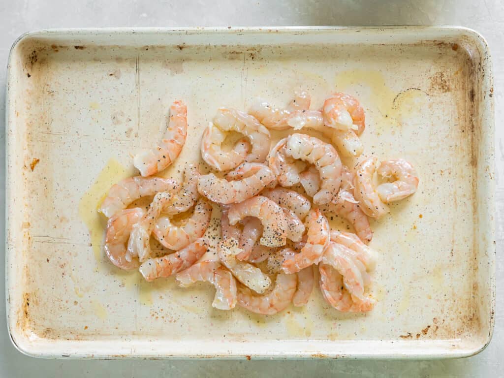 Shrimp drizzled with oil on a baking sheet with a wooden spatula.