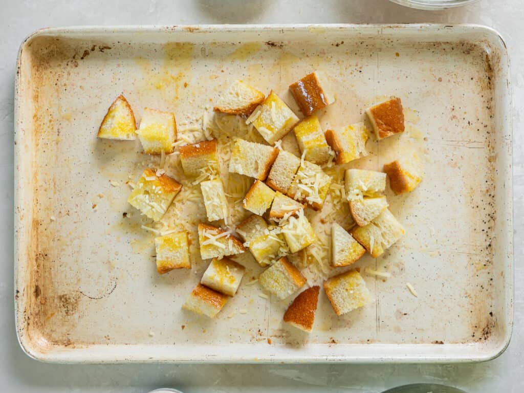 Bread cubes coated with melted butter, oil and Parmesan on a baking sheet.