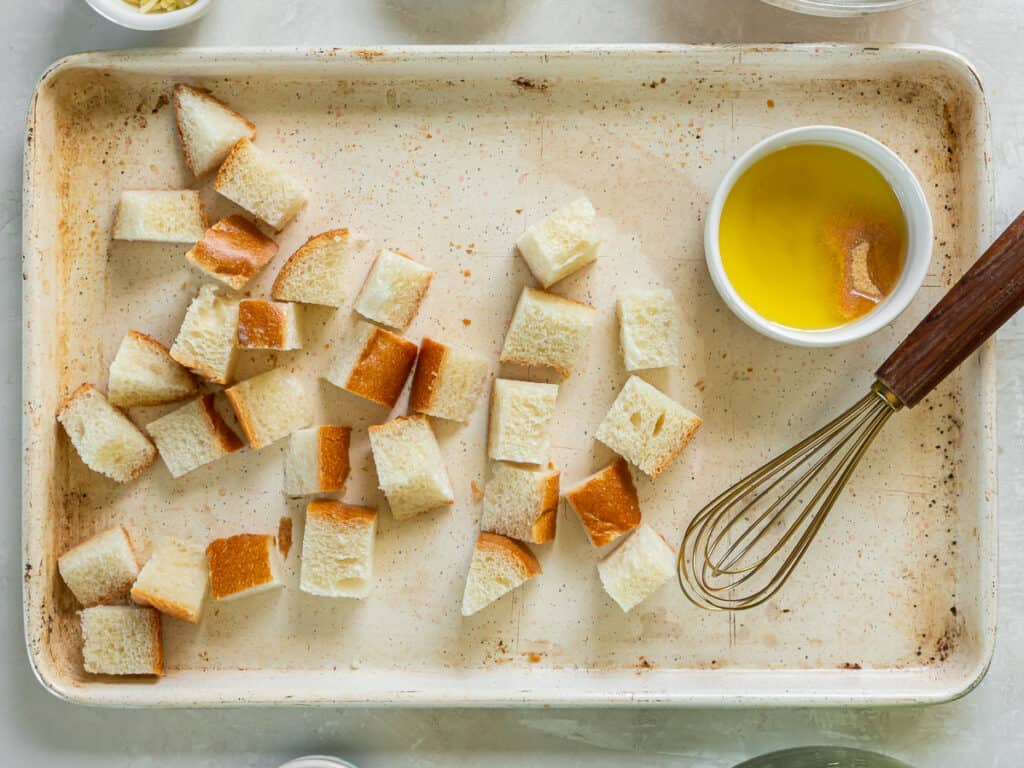 Bread cubes on a baking sheet with a ramekin filled with melted butter and seasoning and a whisk.