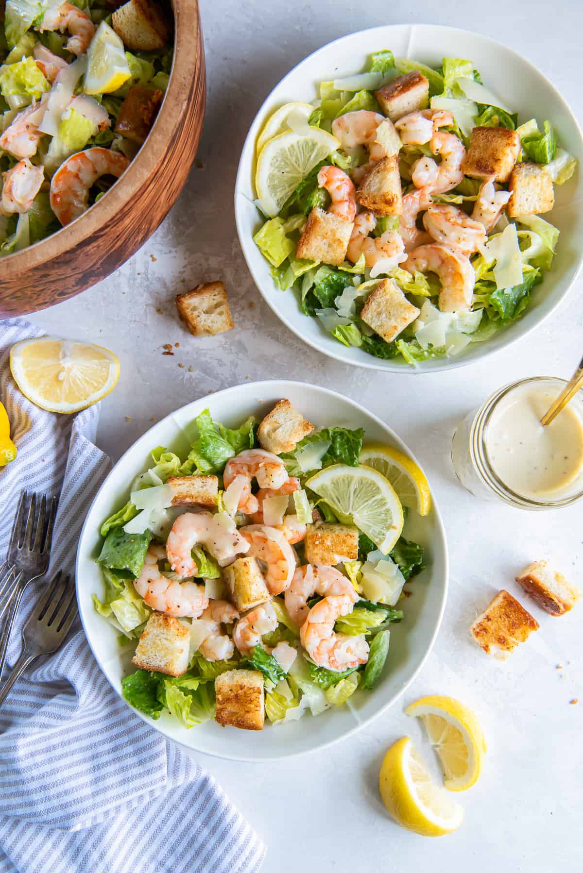 Two bowls of Shrimp Caesar Salad next to a salad bowl and mason jar filled with dressing.
