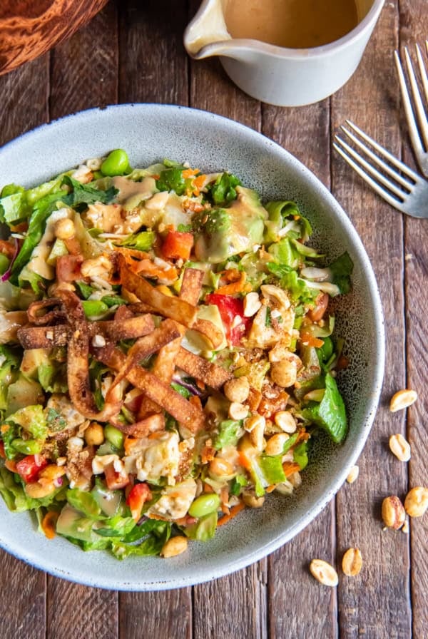 A top down shot of a salad with chicken, edamame, peanut dressing and wonton strips next to a carafe of dressing and forks.
