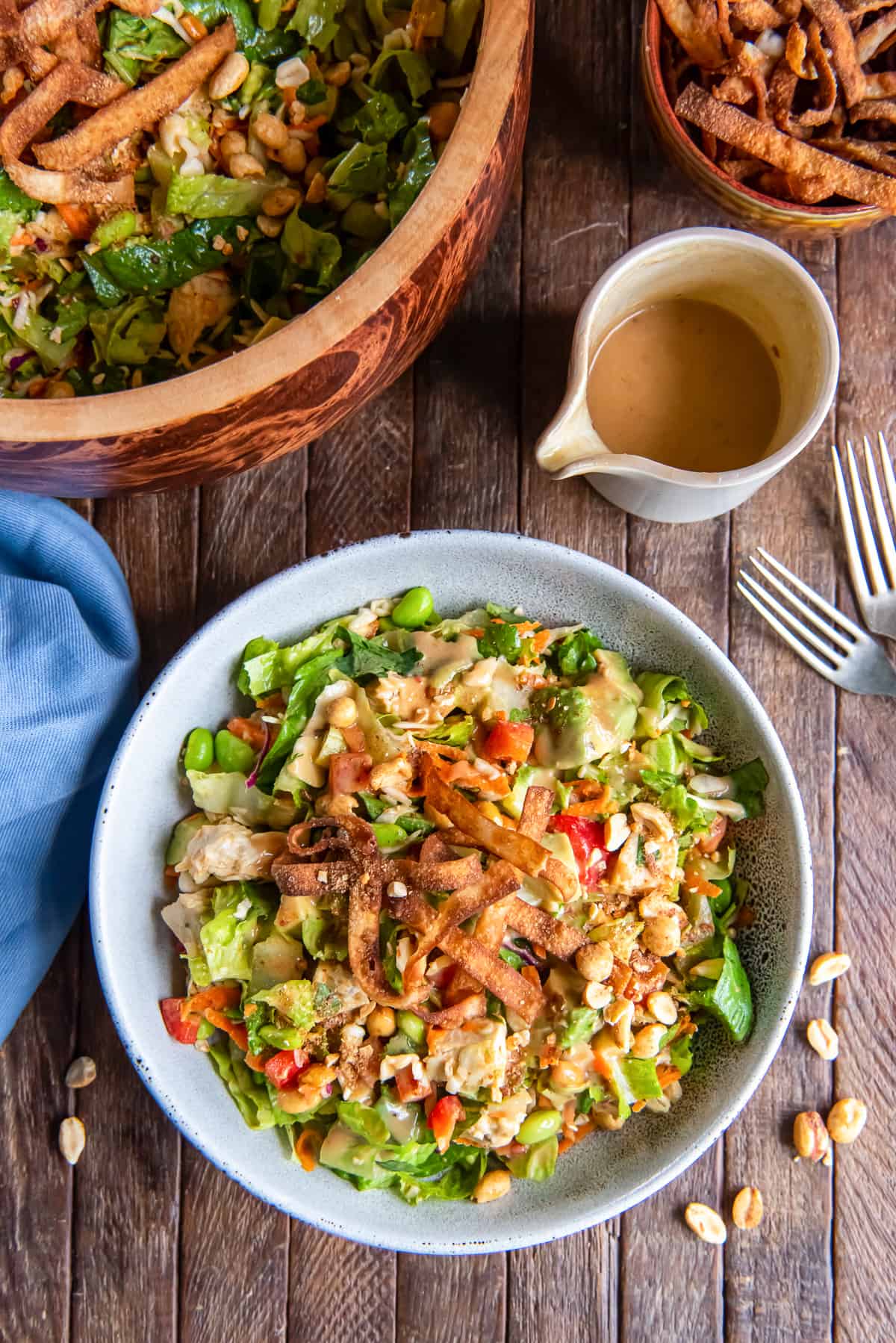 A top down shot of a salad with chicken, edamame, peanut dressing and wonton strips next to a carafe of dressing and forks.