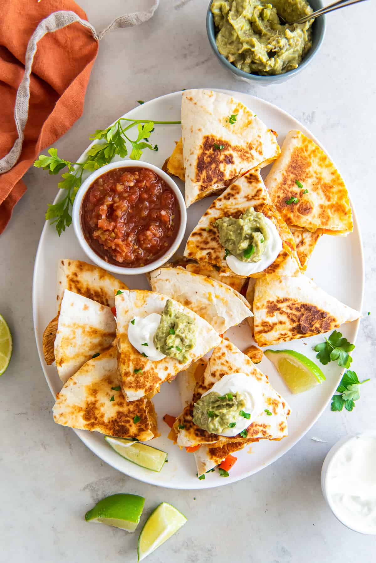 A top down shot of quesadillas cut into wedges on a white platter topped with sour cream and guacamole with a small bowl of salsa.