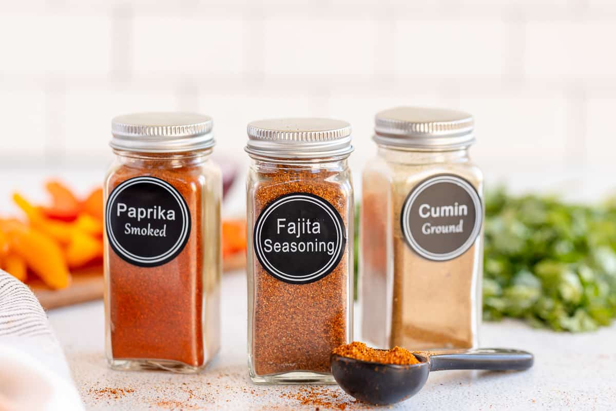Two spice jars on either side of a jar of fajita seasoning with a measuring spoon lying in front.