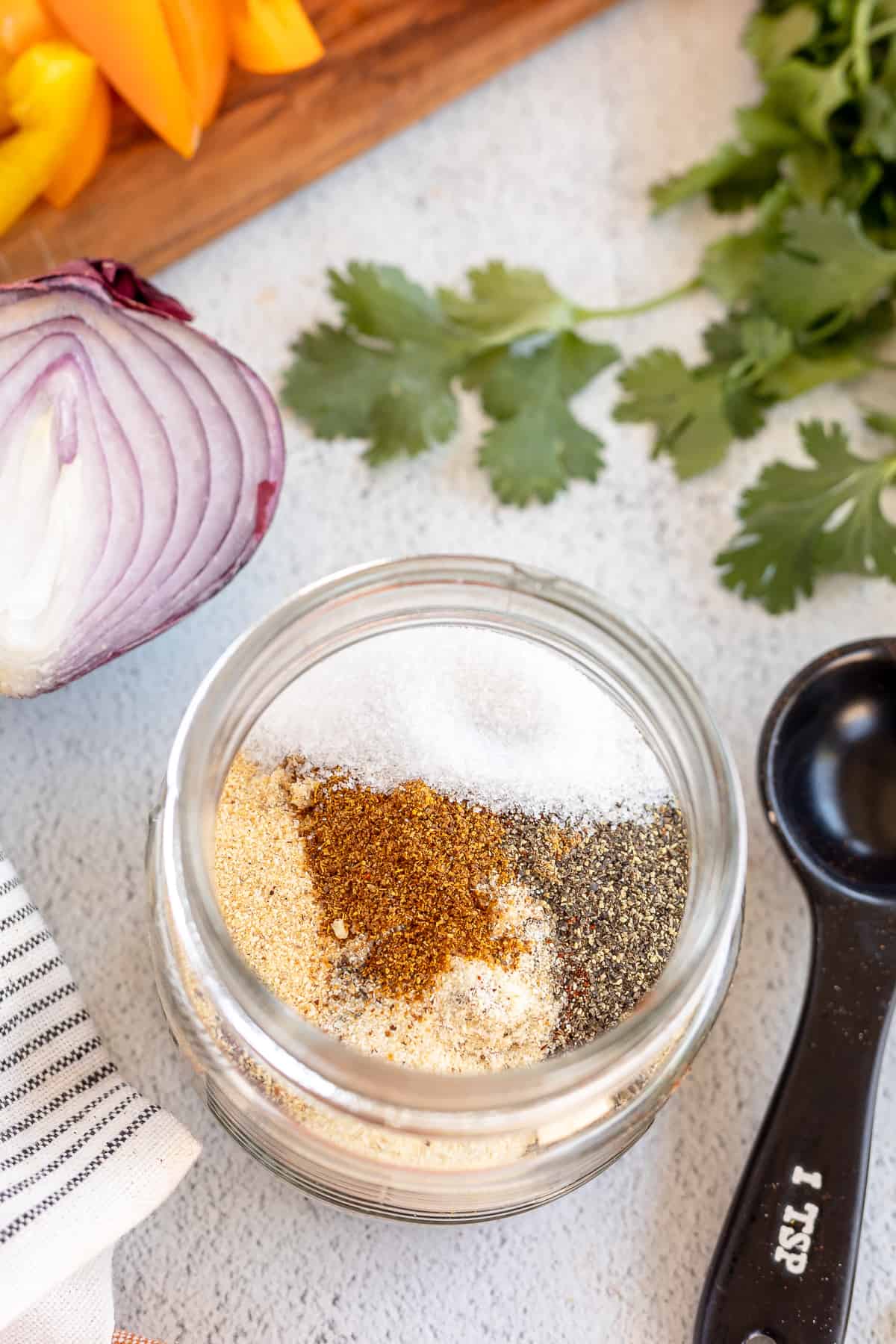 A top down shot of spices in a mason jar next to a red onion, yellow  bell peppers, cilantro, and a measuring spoon.