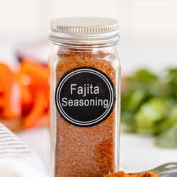 A spice bottle with a label that reads Fajita Seasoning and filled with brown spice next to a measuring spoon full of the spice.