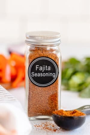 A spice bottle with a label that reads Fajita Seasoning and filled with brown spice next to a measuring spoon full of the spice.