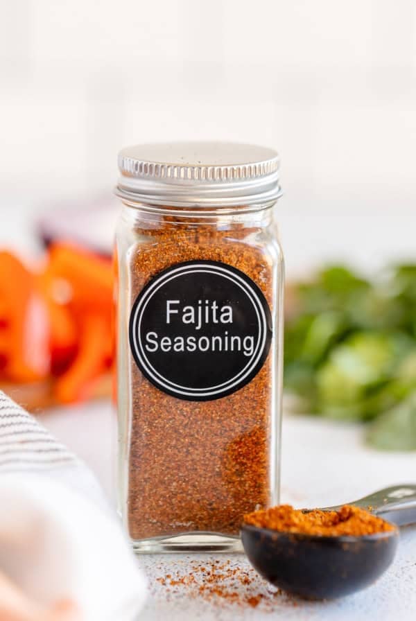 A spice bottle with a label that reads Fajita Seasoning and filled with brown spice next to a measuring spoon full of the spice.