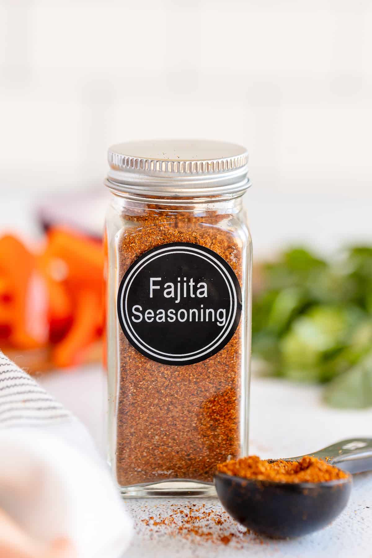 A spice bottle with a label that reads Fajita Seasoning and filled with brown spice next to a measuring spoon full of the spice. 