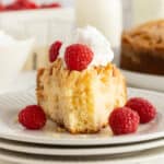 A slice of almond pound cake topped with whipped cream and raspberries with a bite missing on a white plate.