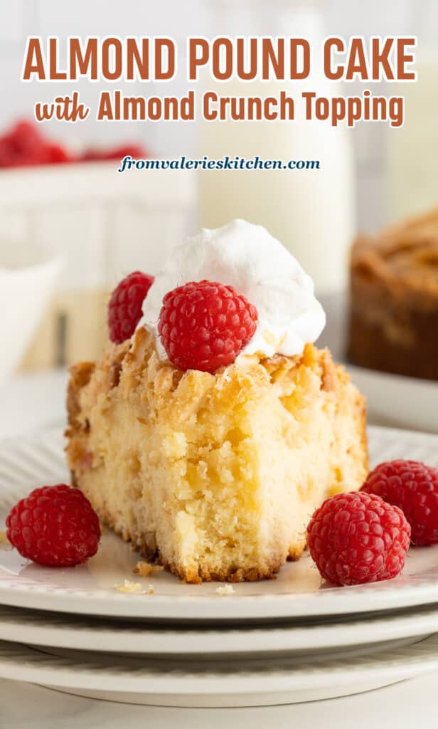 A slice of almond pound cake topped with whipped cream and raspberries with a bite missing on a white plate with text.