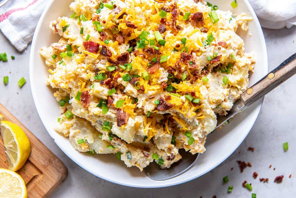 A top down shot of a spoon resting in a bowl of potato salad with cheese and bacon.