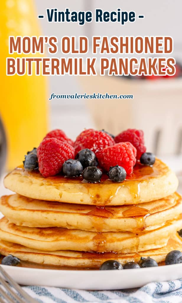 A stack of pancakes topped with fresh berries and maple syrup on a white plate with a glass pitcher of orange juice in the background with text.