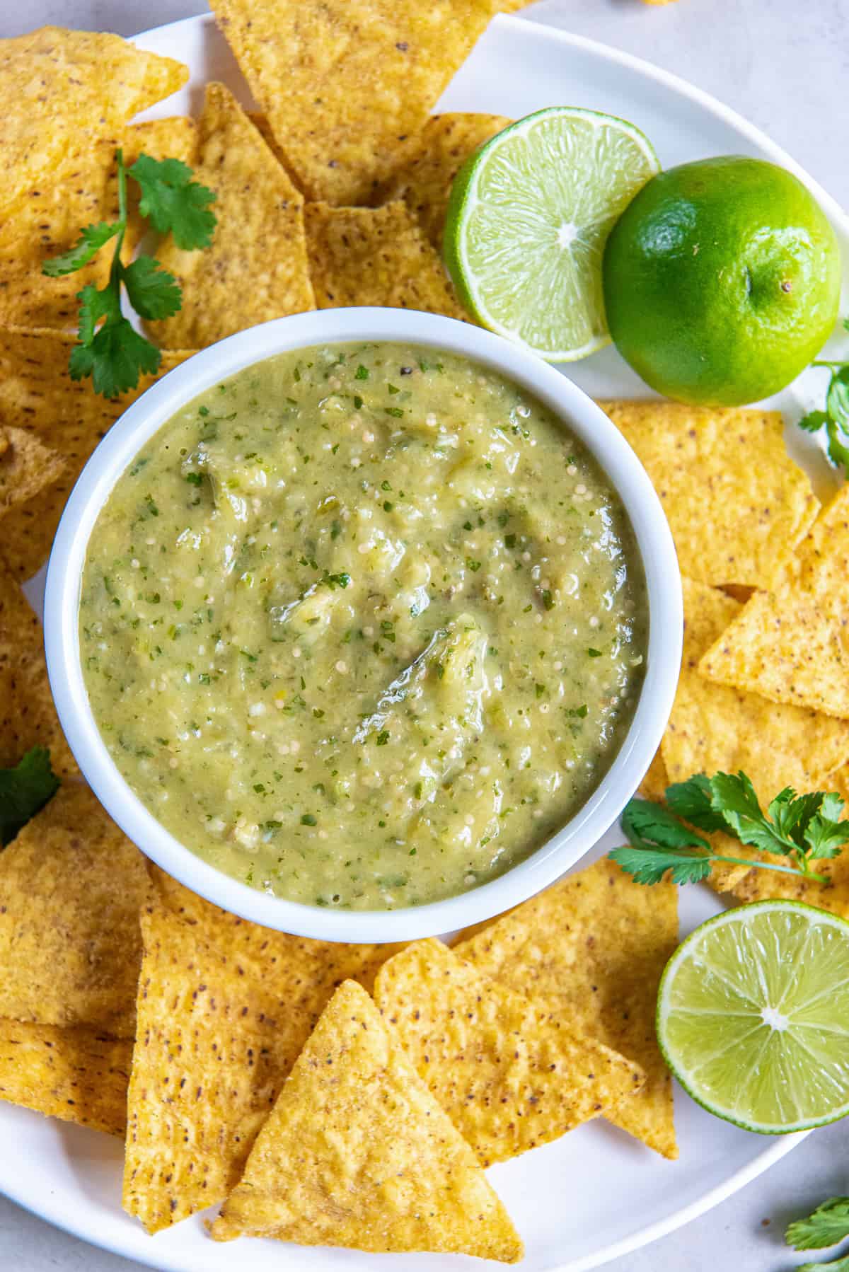 A top down shot of a small bowl of salsa verde on a white plate with tortilla chips.