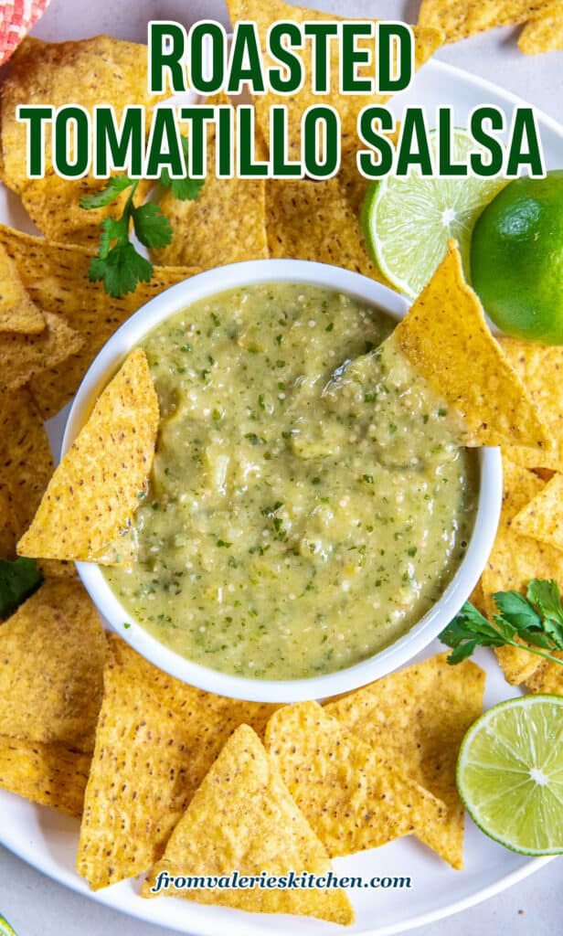 A top down shot of a small bowl of tomatillo salsa on a plate with tortilla chips, lime halves, and sprigs of cilantro with text.